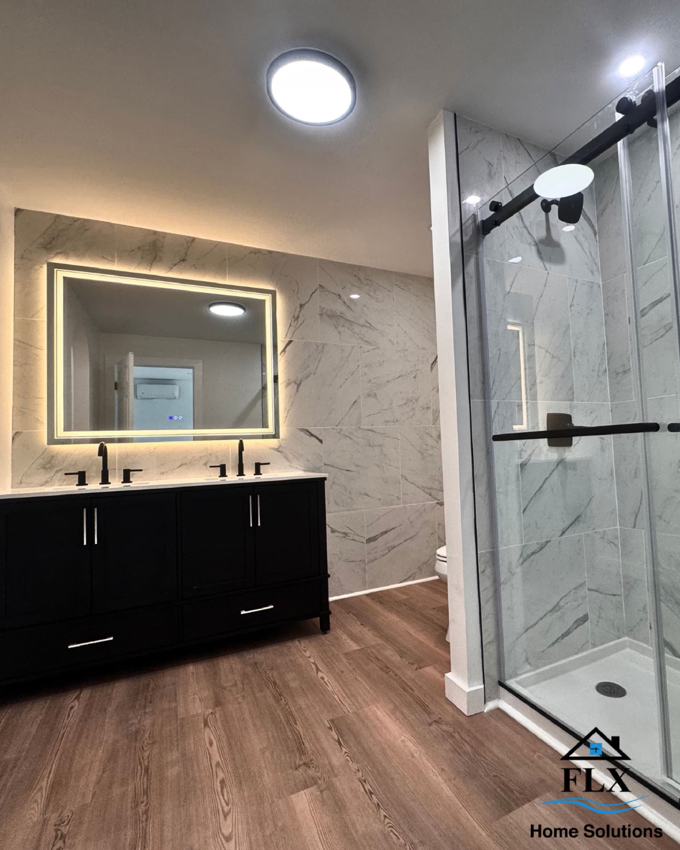 Modern bathroom with black vanity, marble tile walls, glass shower, and wood-look flooring