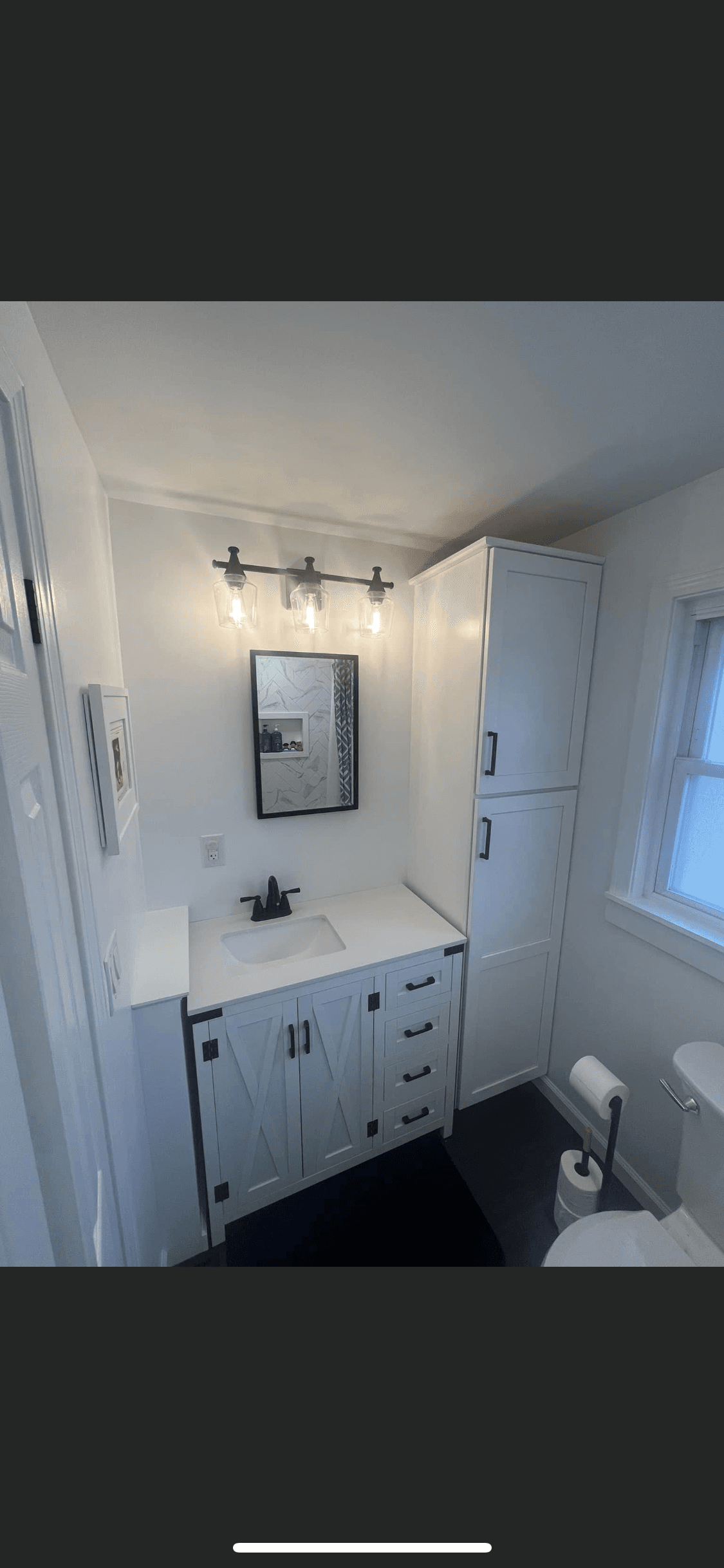 White farmhouse vanity with black fixtures and tall cabinet in remodeled bathroom