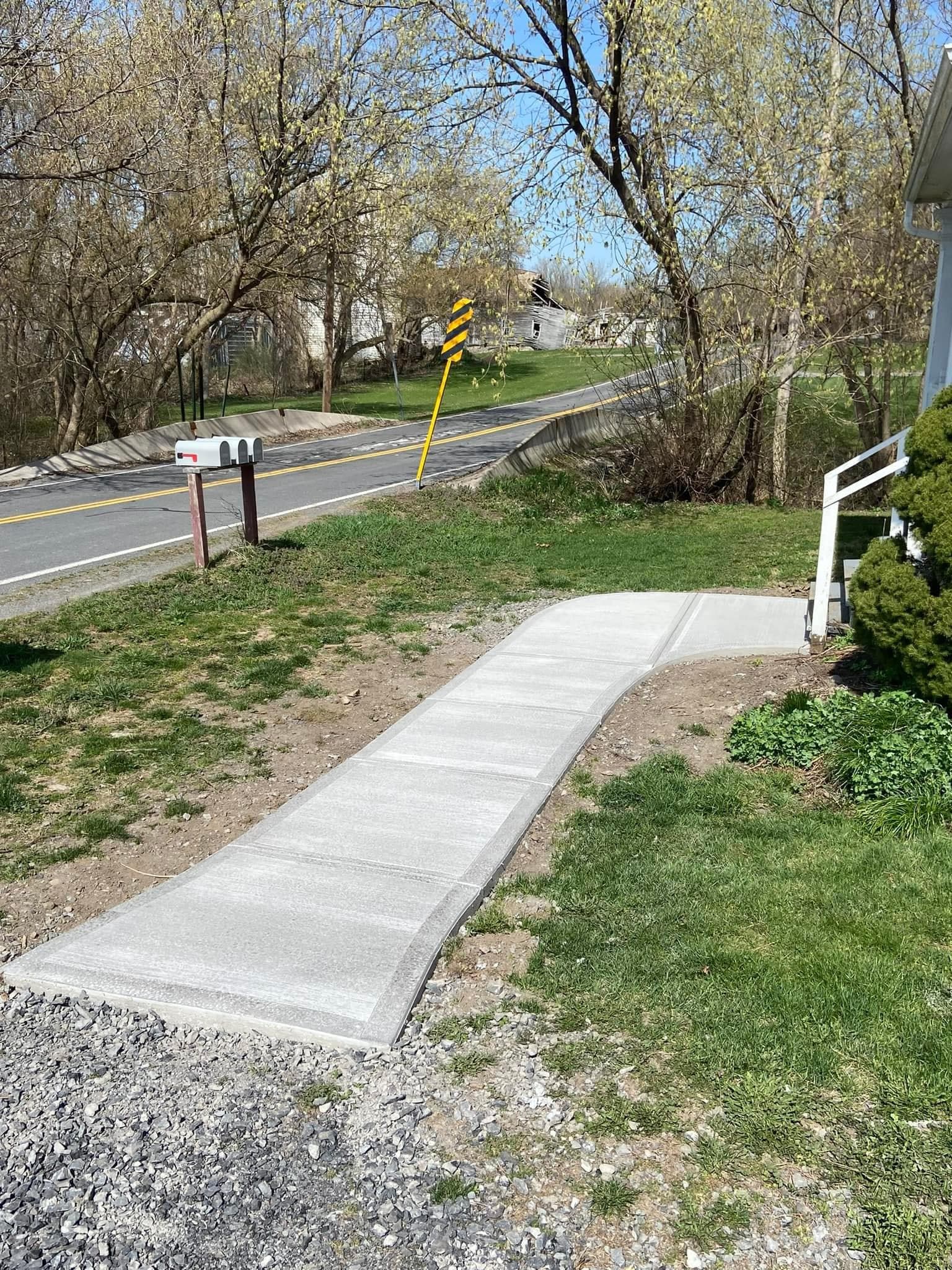 Curved concrete walkway leading from lawn to rural road with yellow chevron sign