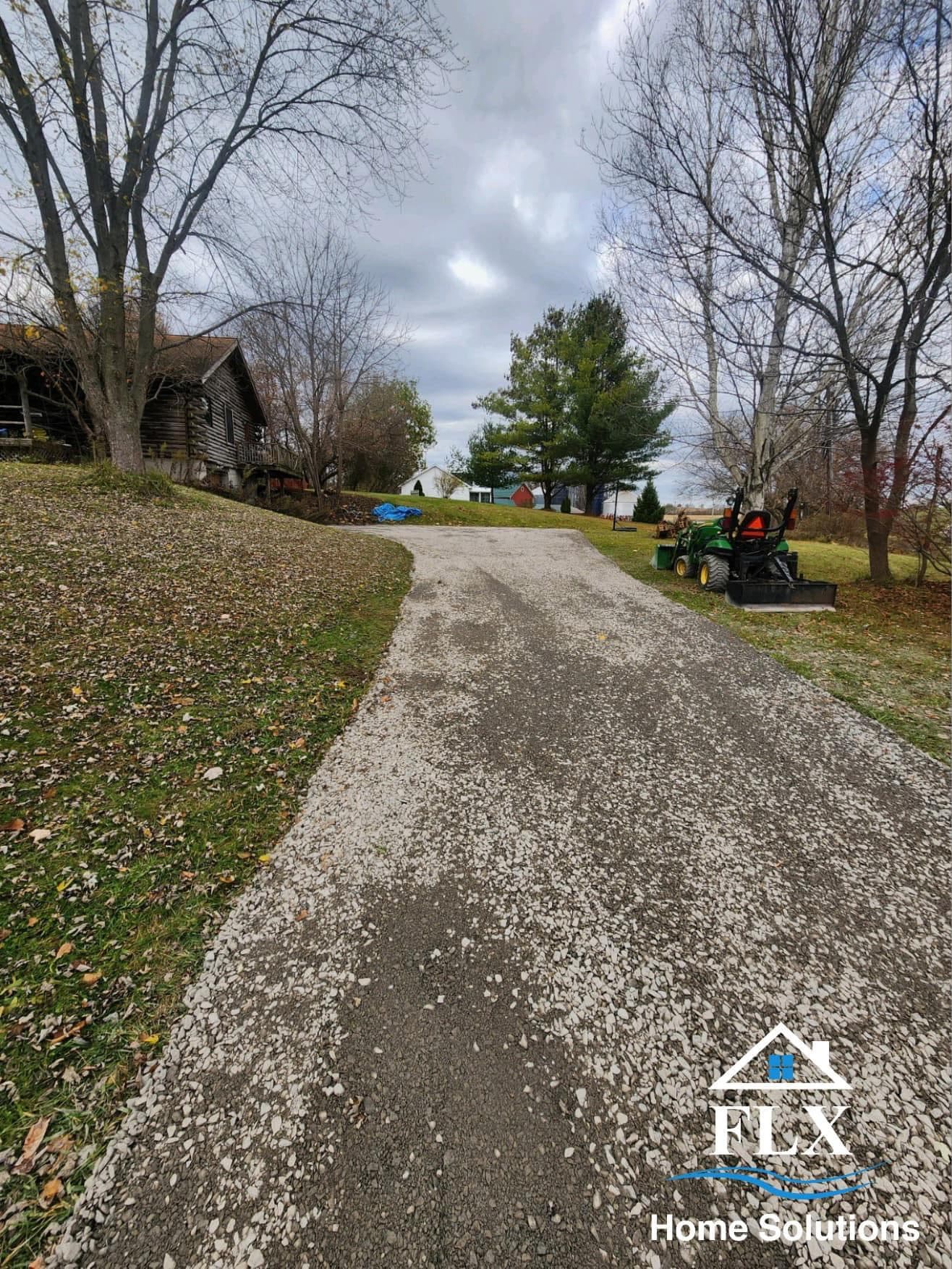 Gravel driveway leading to rustic wooden home with bare trees and green tractor