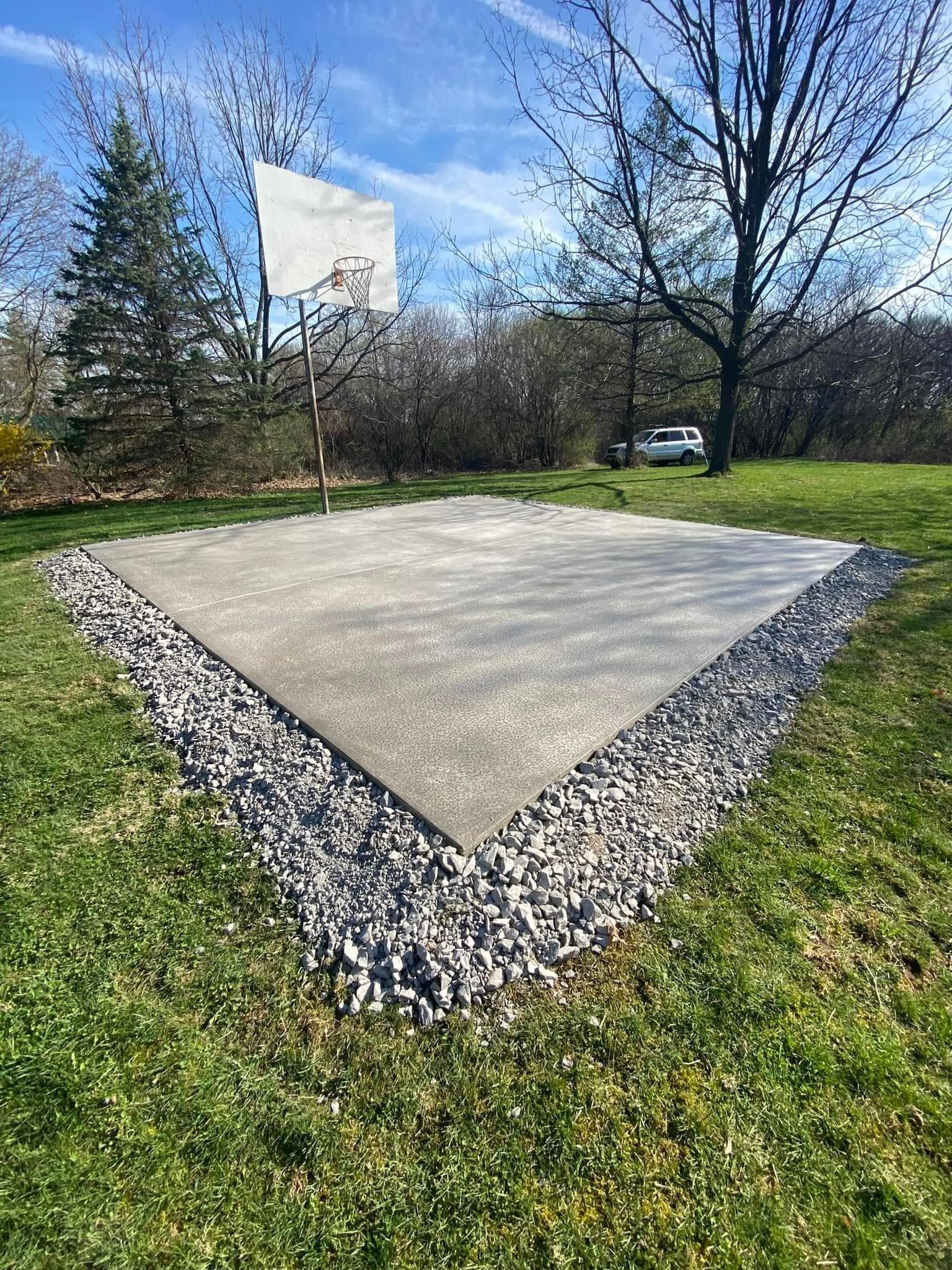 Concrete basketball court pad with white backboard and rock border in backyard