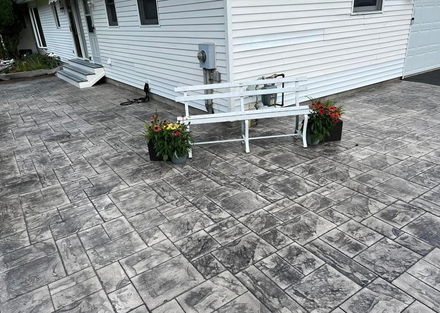 Stamped concrete patio with white bench and flower planters beside white house