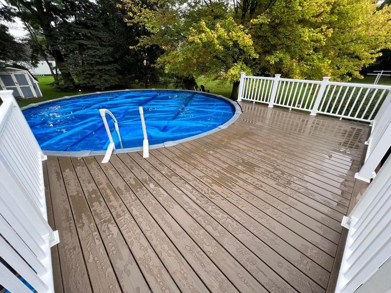 Above ground pool with wet composite deck and white railing surrounded by trees
