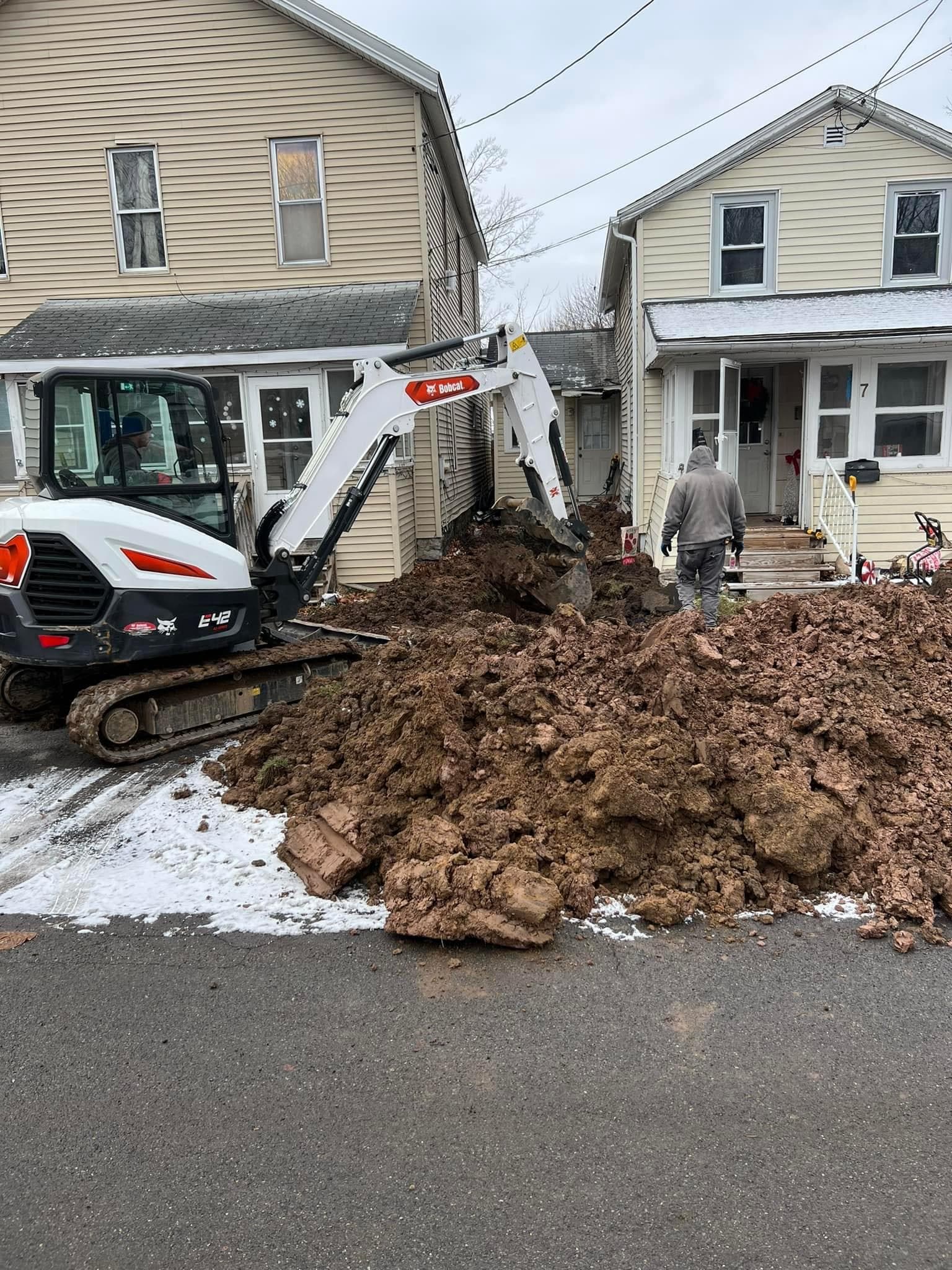 Bobcat excavator digging between two residential houses with large dirt pile on street