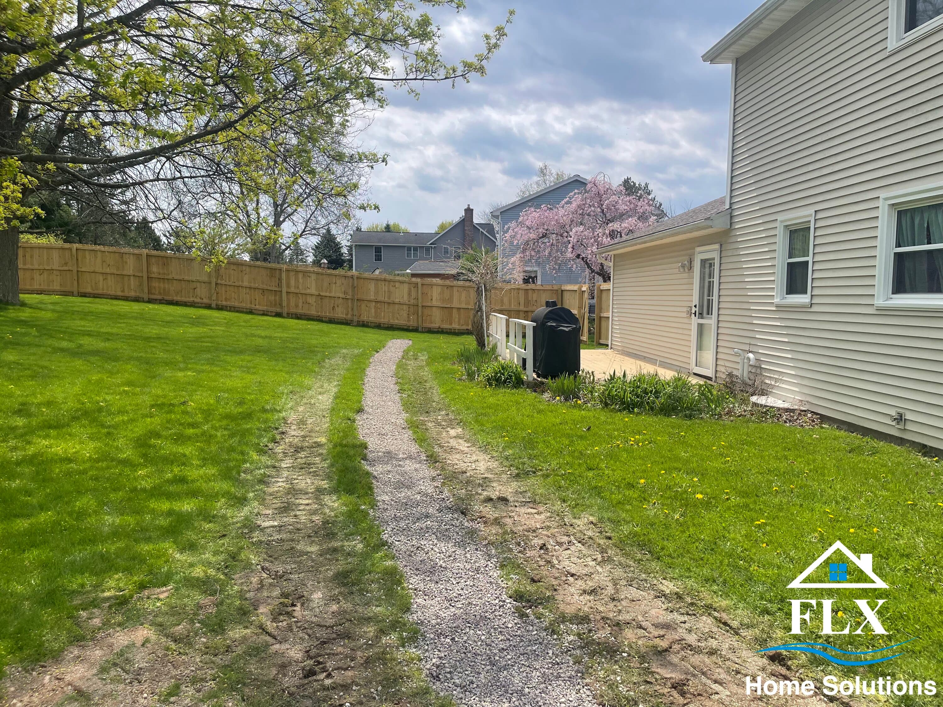 Gravel driveway path through green lawn beside house with wooden fence